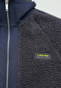 Το φλις μπουφάν Calvin Klein διαθέτει σκούρο ναυτικό χρώμα, υλικό με υφή χνουδωτή, μια λεία υφασμάτινη επιφάνεια και μια διακριτή ετικέτα με το λογότυπο.