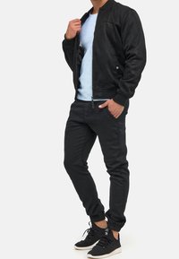 Zwarte suedine bomberjack met ritssluiting, gedragen over een lichtblauw shirt, gecombineerd met zwarte tapered broeken en zwarte sneakers.