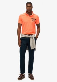 Jasnopomarańczowa koszulka polo z czarnym napisem "Super Dry Athletics", szary sweter związany w talii, granatowe spodnie, brązowe buty na sznurówki.