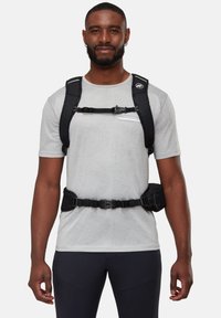 Svart ryggsäck med vadderade remmar, justerbara bröst- och midjebälten och en texturerad yta, bärs över en grå kortärmad skjorta.