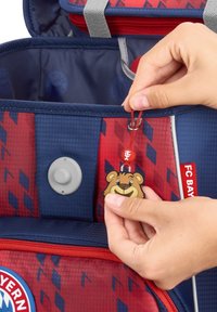 Sac à dos du Bayern Munich avec des motifs géométriques rouges et bleus, du matériel gris et un pendentif en forme d'ours attaché à une corde rouge.