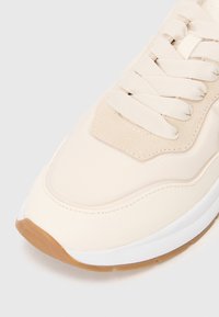 Közelkép egy bézs sneaker-ről, amely szövet és velúr panelekből készült, krém színű fűzőkkel, fehér középtalppal és gumitalppal, fehér háttéren.