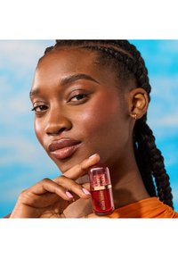 Botella de rubor rojo con tapa redondeada, acentos dorados, sostenida por una persona. Tono de piel medio, pelo trenzado, vistiendo una blusa naranja brillante.