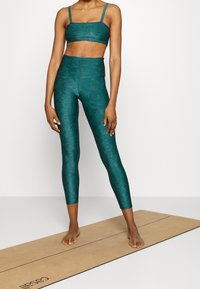 Akvamarin sportig crop top och leggings, texturerade med ett subtilt mönster. Materialet verkar vara stretchigt. Modellen står barfota på en yogamatta.
