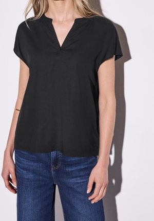 Kobieta ubrana w luźną czarną bluzkę z krótkim rękawem, z rozciętym dekoltem w kształcie litery V, oraz niebieskie dżinsy, stojąca na białym tle.