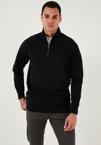 Melna kapucīna džemperis ar pus-izsietu apkakli, raglan piedurknēm un minimālu logotipa detaļu. Mīksta tekstūra un pieguļošs dizains.