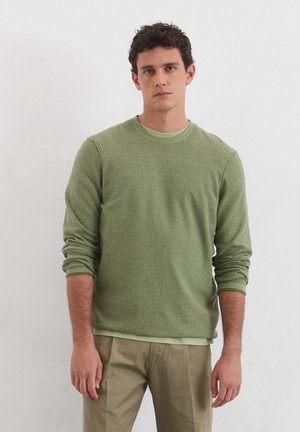Grüner, langärmliger Pullover mit strukturiertem Finish, Rundhalsausschnitt und gerippten Bündchen, kombiniert mit hellbeigen Hosen.