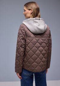 Frau blickt nach links, trägt eine braune Steppjacke mit einer hellgrauen Kapuze und blaue Jeans vor einem grauen Hintergrund.