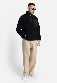 Sudadera de forro polar negra con media cremallera y bolsillo frontal, combinada con pantalones rectos beige y zapatillas gris claro. El modelo lleva gafas de sol redondas.