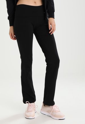 Femme portant un pantalon noir ajusté, une veste courte et des chaussures de sport rose clair, debout devant un fond clair uni.