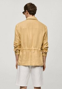 Hořčicově zbarvená lněná košile s uvolněným střihem, zapínáním na manžetách a nařaseným detailem v pase, spárovaná s bílými šortkami.
