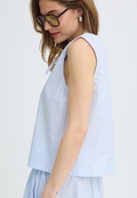 Femme portant un haut sans manches à rayures verticales bleues et blanches et une jupe assortie, avec des cheveux bruns, une boucle d’oreille en argent et de grandes lunettes de soleil.