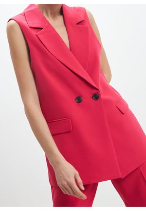 Femme portant un blazer sans manches rouge vif à double boutonnage avec des boutons noirs et un pantalon rouge assorti sur fond blanc.