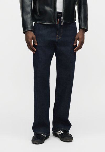 Άνδρας που φοράει σκούρο μπλε τζιν με ίσια γραμμή, μαύρο δερμάτινο μπουφάν, μαύρα αθλητικά παπούτσια με άσπρες ριγέ κάλτσες, στέκεται μπροστά σε λευκό φόντο.