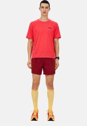 Red sports t-shirt, maroon shorts, yellow knee-high socks, and orange sneakers. Material appears lightweight and breathable, with logo accents.