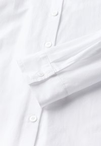 Chemise blanche à manches longues avec des poignets à boutons et des boutons à l'avant, présentée pliée sur une surface plane.