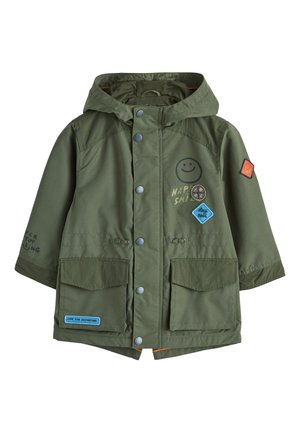 Ungt barn iført en olivengrøn hættejakke med lapper, sorte bukser og sorte sneakers, stående og kigger let til siden.