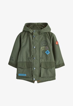 Niño pequeño con un abrigo verde oliva con capucha y parches, pantalones negros y zapatillas negras, de pie y mirando ligeramente hacia un lado.
