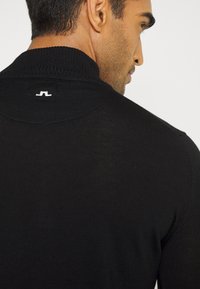 Black ribbed sweater featuring a high collar and a small white logo at the back, showcasing a fitted design and textured fabric.