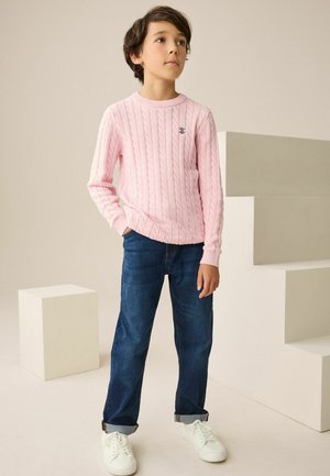 Rosa kabelstrikket sweater med rund hals, kombineret med blå jeans og hvide sneakers. Barn står på en lys baggrund med geometriske former.