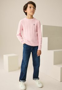 Rožinis megztinis su pynimo raštu ir apvalia iškirpte, derinamas su mėlynais džinsais ir baltais sportbačiais. Vaikas stovi ant šviesios fono su geometrinėmis formomis.