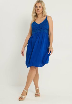 Kobieta ubrana w bezrękawnikową sukienkę w kolorze królewskiego błękitu z koronkowym gorsetem oraz złote sandały na koturnie, stojąca na prostym jasnym tle.