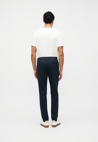 Homme debout de dos portant un t-shirt blanc, un pantalon à taille élastique bleu marine et des baskets blanches sur un fond uni.