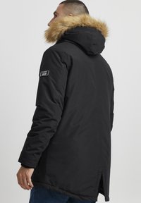 Sort parka-jakke med en faux fur hætte, mærke på venstre ærme og lige snit med sideslidser i kanten.