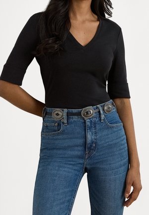 Kvinde iført en sort V-hals top med albuelange ærmer og blå højtaljede jeans med et bredt dekorativt bælte.