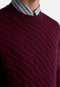 Bordeauxroter Strickpullover mit strukturiertem Zopfmuster, mit Rundhalsausschnitt und über einem gestreiften Hemd mit blau-weißen Streifen getragen.