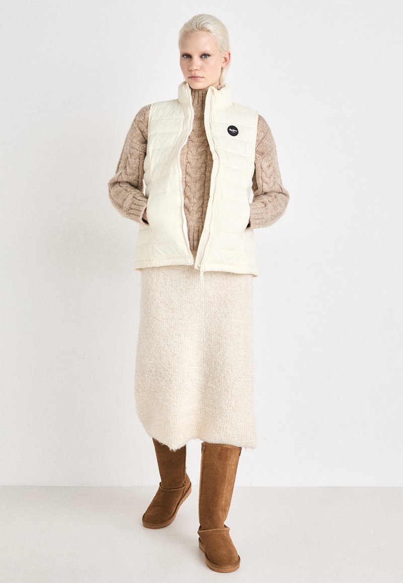 Gilet rembourré blanc sur un pull en maille torsadée beige, assorti à une jupe en peluche beige et des bottes en suède marron. Comprend un écusson noir.