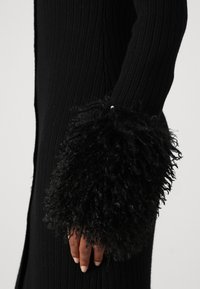 Zwarte geribbelde trui met brede, gestructureerde, pluizige manchetten. De stof heeft een zachte, lineaire patroon met een getailleerde silhouet.