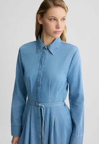 Helles blaues Denimkleid mit langen Ärmeln, Knopfleiste vorne und spitzem Kragen. Mit einem taillierten Gürtel und einem gefältelten Rock für eine strukturierte Form.