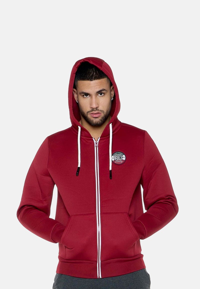 Homme portant un sweat à capuche rouge zippé avec des cordons blancs et des poches avant, debout les mains dans les poches devant un fond uni.