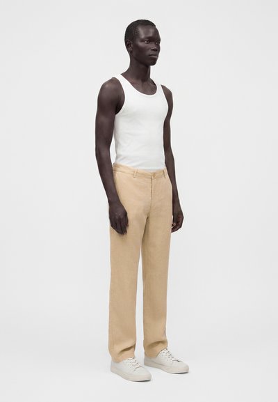 Άνδρας που στέκεται φορώντας λευκό αμάνικο πουκάμισο, μπεζ παντελόνι και λευκά αθλητικά παπούτσια σε ένα απλό ανοιχτό φόντο.