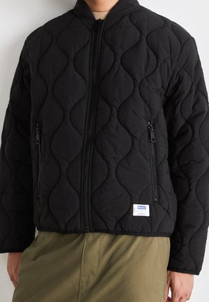 Zwarte doorgestikte jas met ritssluiting en capuchon, twee ritssluitzakken en een klein wit rechthoekig merkje op de voorkant van de zoom.