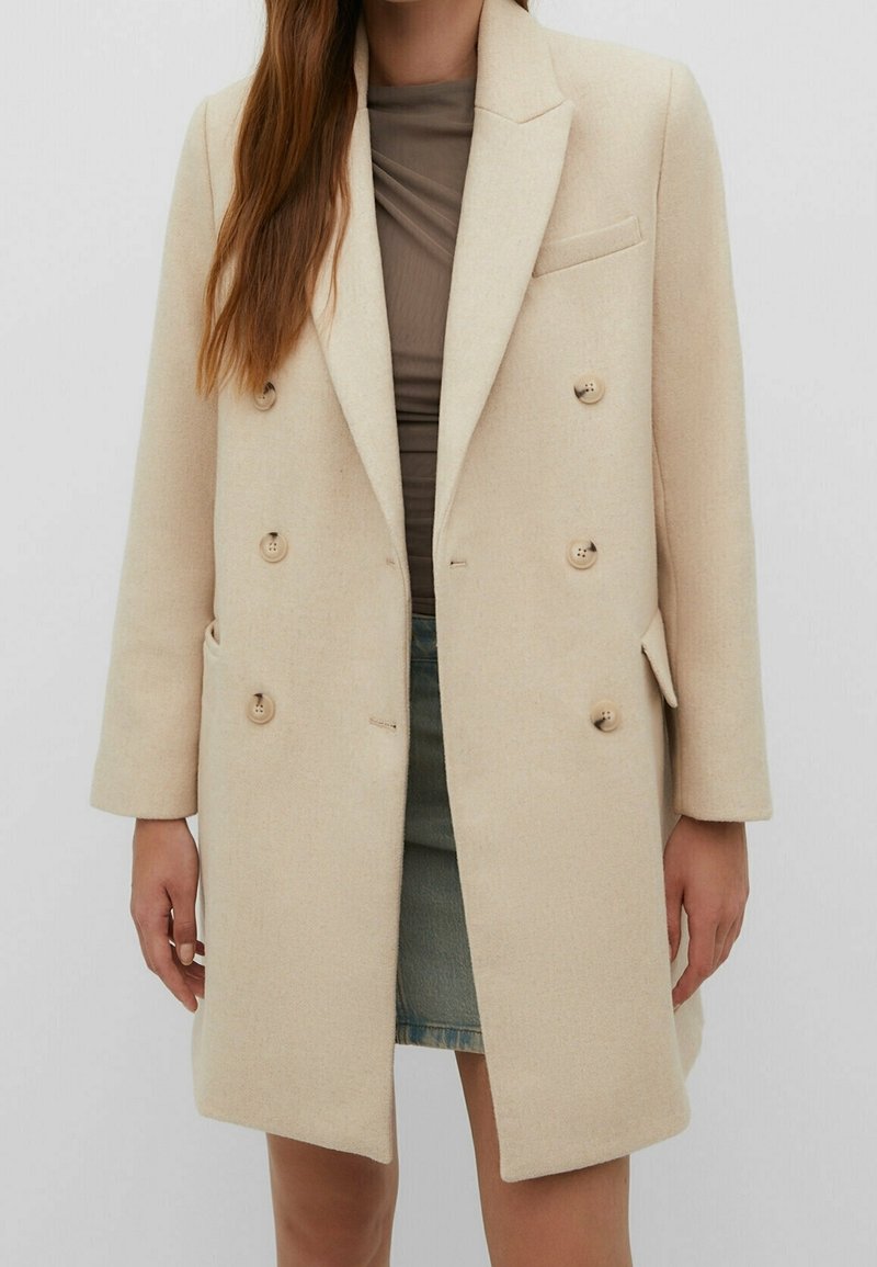 Femme portant un manteau en laine beige à double boutonnage sur un top marron et une jupe en jean bleue, sur un fond uni.