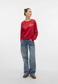 Sweat-shirt en coton rouge avec le texte "ÉTABLI EN 1986", associé à un jean en denim bleu clair à jambes larges et des chaussures marron. Design simple, style décontracté.