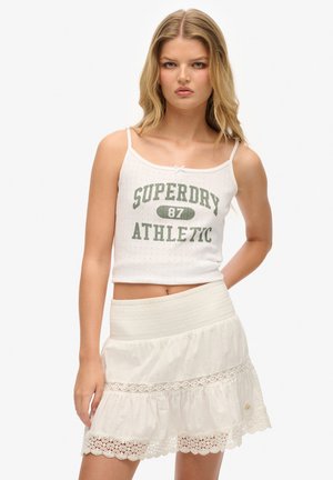 Weißes Tanktop mit dem Aufdruck "SUPERDRY 87 ATHLETIC" in Grün, kombiniert mit einem weißen, ausgestellt geschnittenen Rock mit Spitze und strukturiertem Stoff.