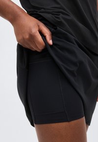 Χέρι σηκώνει μαύρο πουκάμισο για να αποκαλύψει εφαρμοστά μαύρα αθλητικά σορτς με πλαϊνή τσέπη στον μηρό και το ισχίο ενός ατόμου.