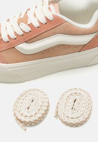 Pink suede sneaker with white accents and a textured sole, accompanied by round cotton shoelaces in white with a peach checkered pattern.