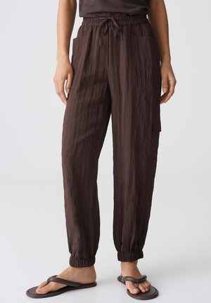 Loose bruine broek met trekkoord, elastische boorden en zijzakken, gedragen met donkere slippers op een lichte achtergrond.