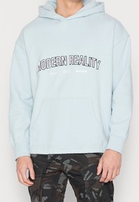 Světlomodrá mikina s kapucí vybavená přední kapsou ve tvaru klokánka a nápisem „MODERNÍ REALITA“. Měkká látka s žebrovanými manžetami a lemem.