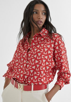 Camicia rossa con bottoni, caratterizzata da un motivo geometrico bianco e crema. Presenta un colletto e maniche arrotolate. Abbinata a una cintura coordinata e pantaloni crema.