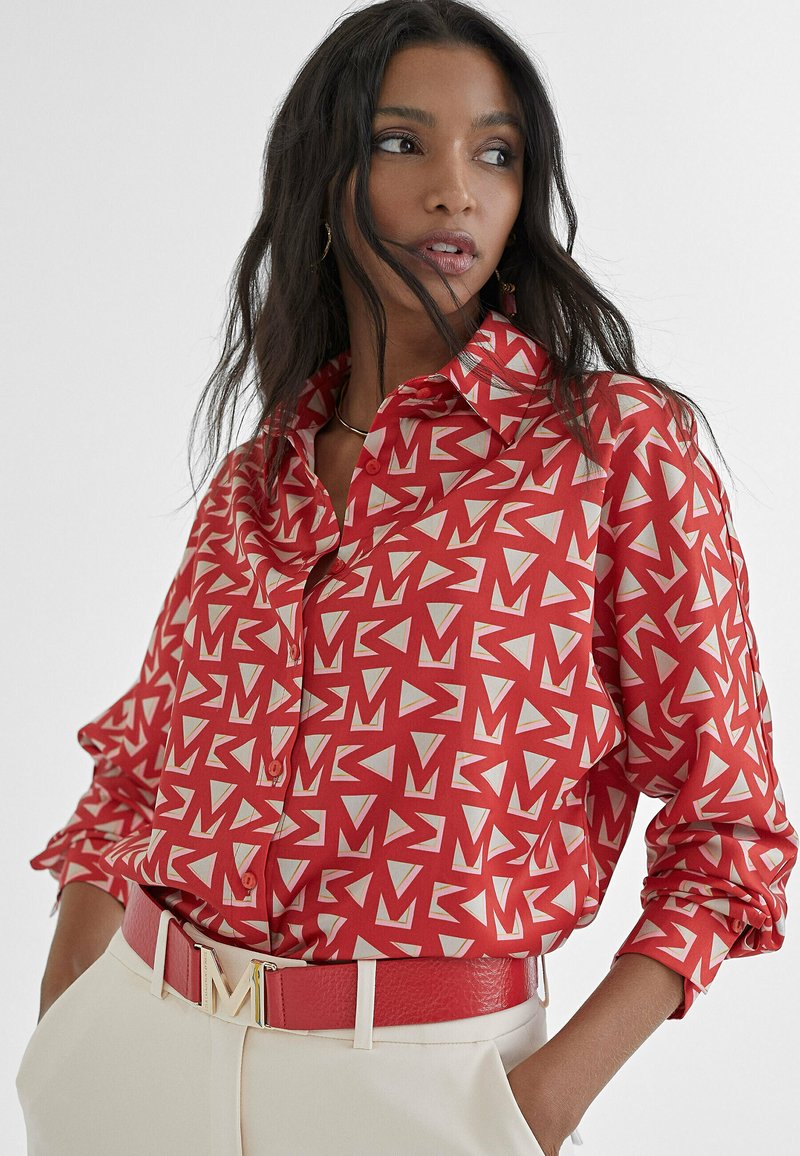 Camicia rossa con bottoni, caratterizzata da un motivo geometrico bianco e crema. Presenta un colletto e maniche arrotolate. Abbinata a una cintura coordinata e pantaloni crema.
