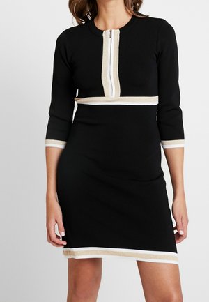 Schwarzes knielanges Kleid mit Dreiviertelärmeln, beigen und weißen Zierakzenten am Reißverschluss, in der Taille, an Ärmelsäumen und Saum.