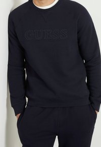 Man med svart sweatshirt och matchande svarta byxor, händerna i fickorna, med en subtilt prägling av "GUESS"-logotypen på framsidan av sweatshirten.