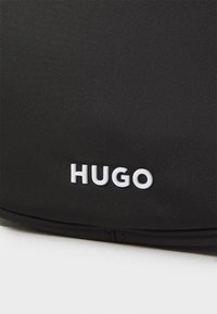 Borsa nera in nylon con superficie testurizzata. La parte anteriore presenta un logo "HUGO" bianco in lettere in grassetto. Chiusura a zip nella parte inferiore.
