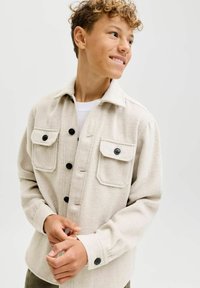 Adolescente con capelli ricci che guarda a destra, indossando una giacca camicia beige con bottoni e due tasche sul petto, e una camicia bianca sotto.