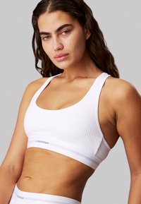 White ribbed bralette with a wide elastic band featuring a subtle logo. Smooth texture and sporty design with thin straps.
